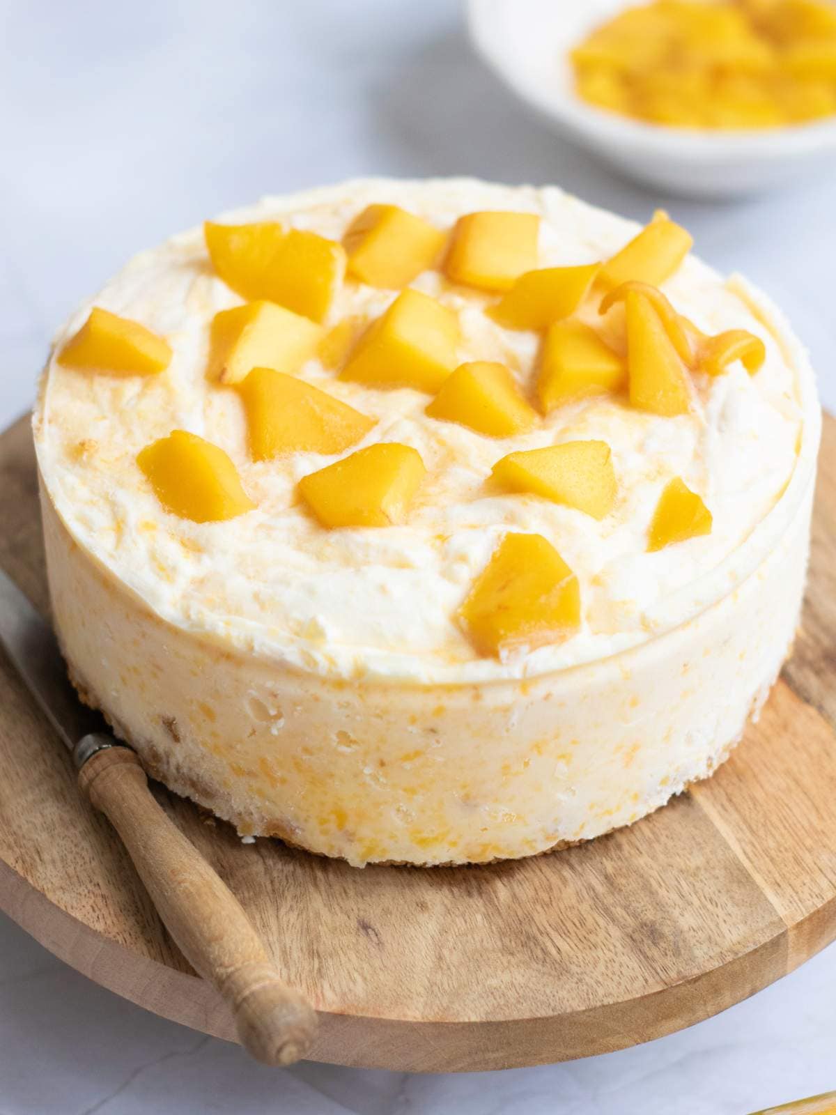 A round cheesecake topped with mango chunks, placed on a wooden board with a knife beside it.
