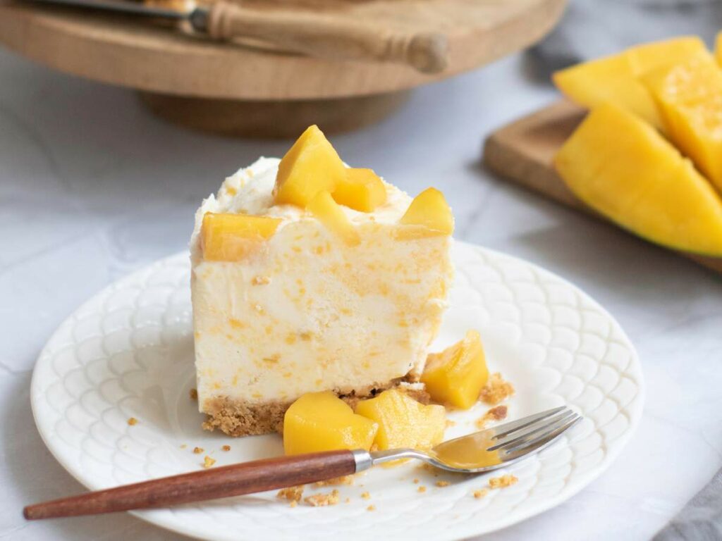 A white plate of cheesecake topped with mango chunks sits on a white plate with a fork, next to sliced mango pieces on a cutting board.