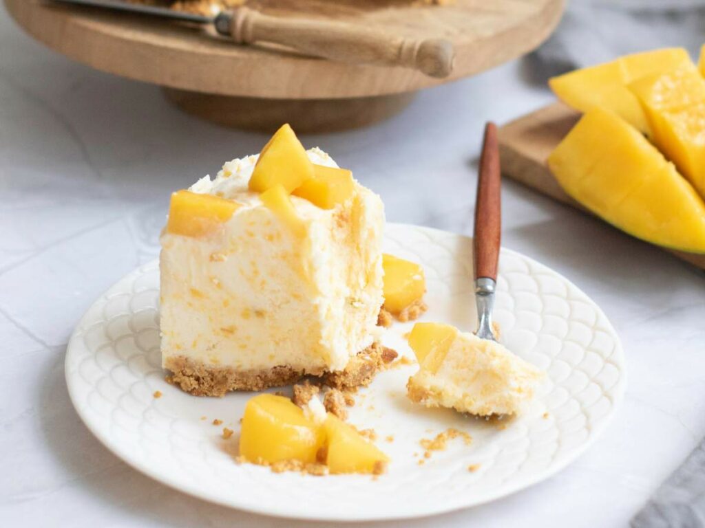 A slice of cheesecake topped with mango pieces sits on a white plate with a fork.