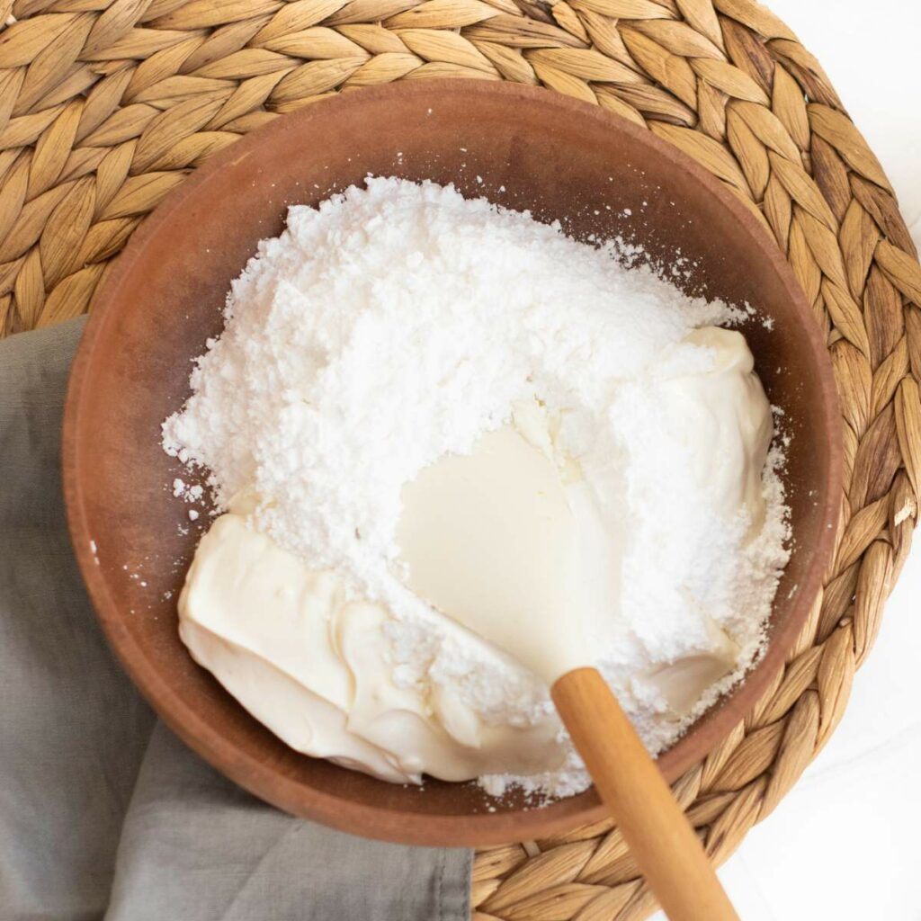 A brown bowl with cream cheese and powdered sugar being mixed with a spatula.