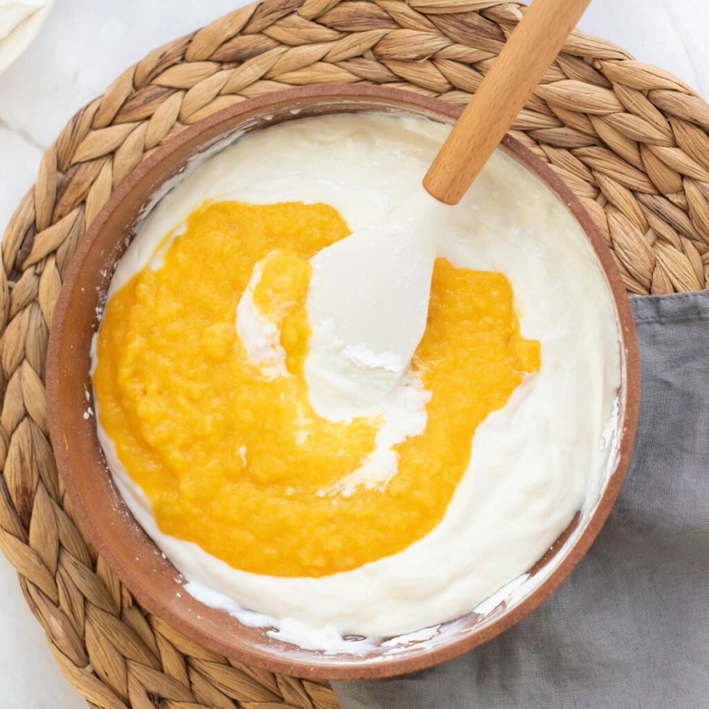 A bowl with a mixture of mango puree and white cream being stirred.