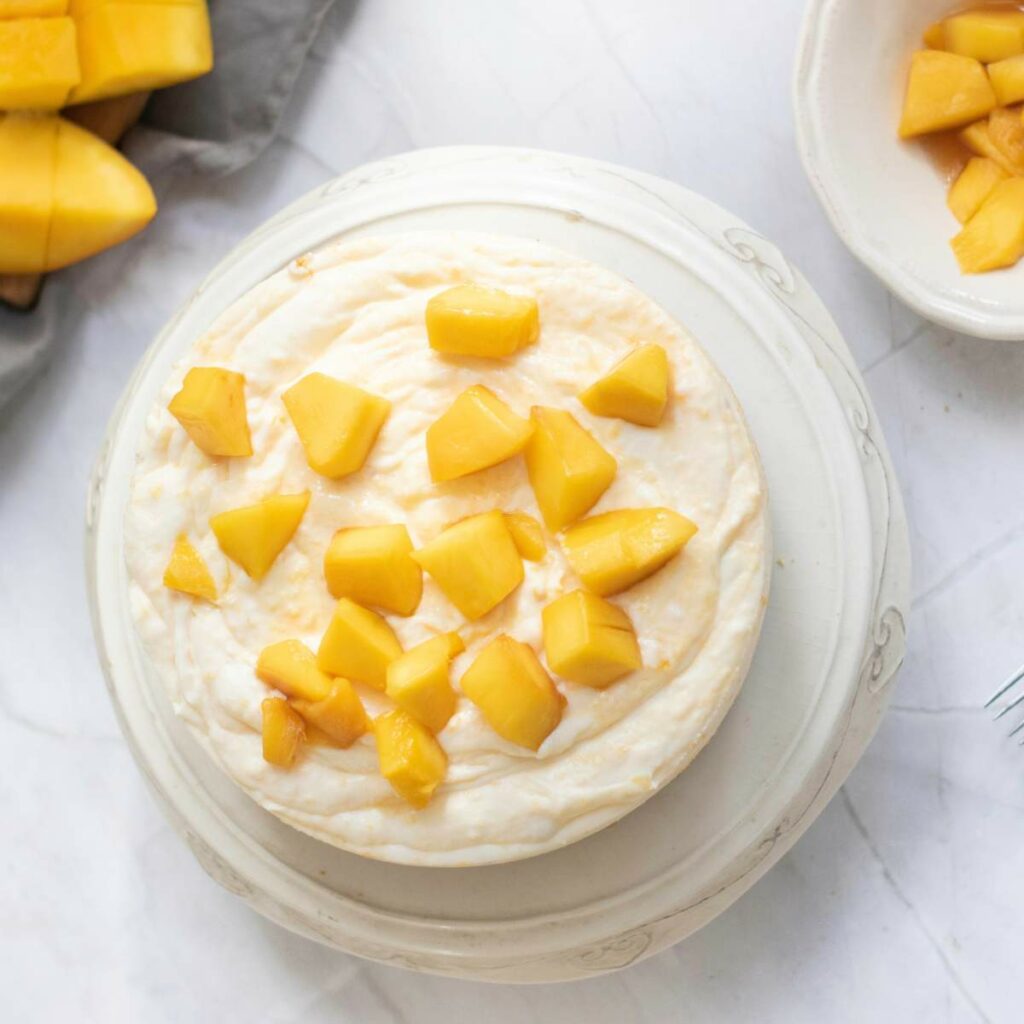 A round cheesecake topped with diced mango pieces sits on a white plate.