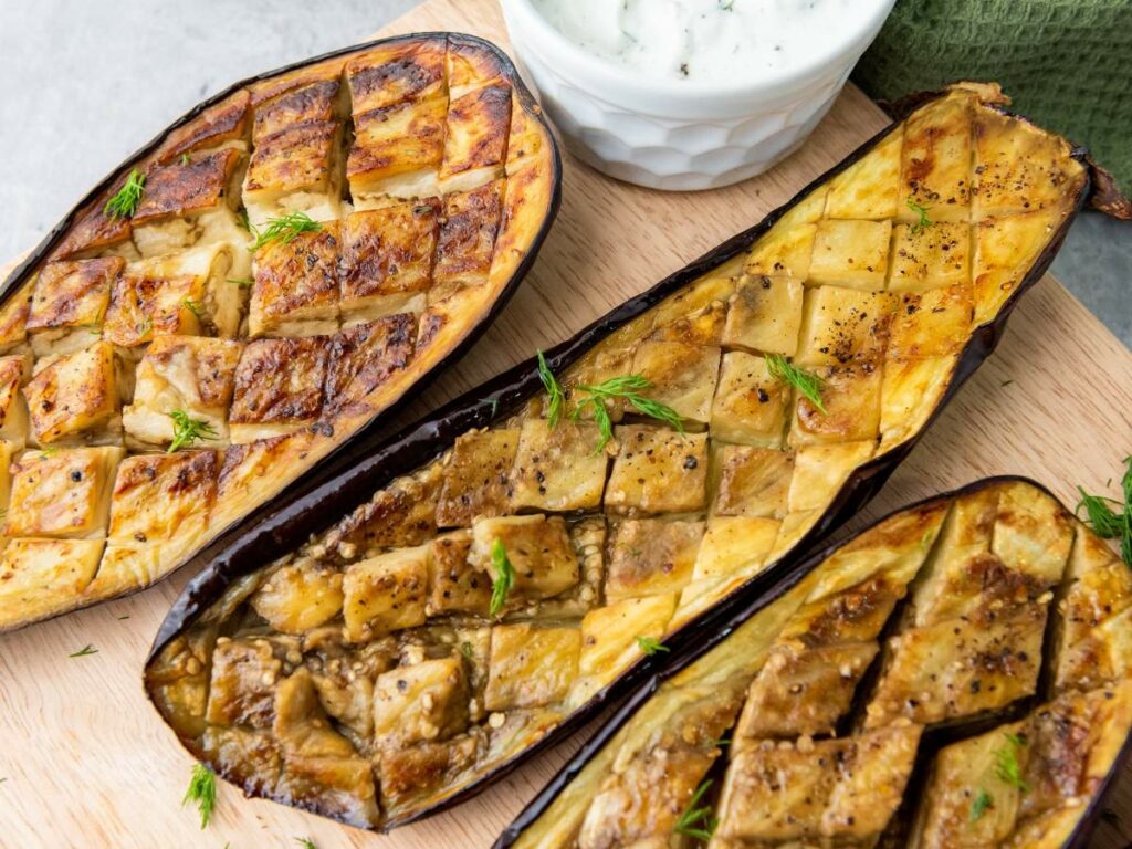 Three roasted eggplant halves with scored, garnished with herbs, next to a small bowl of white sauce.