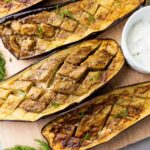 Four roasted eggplant halves with a scored pattern are arranged on a wooden board.