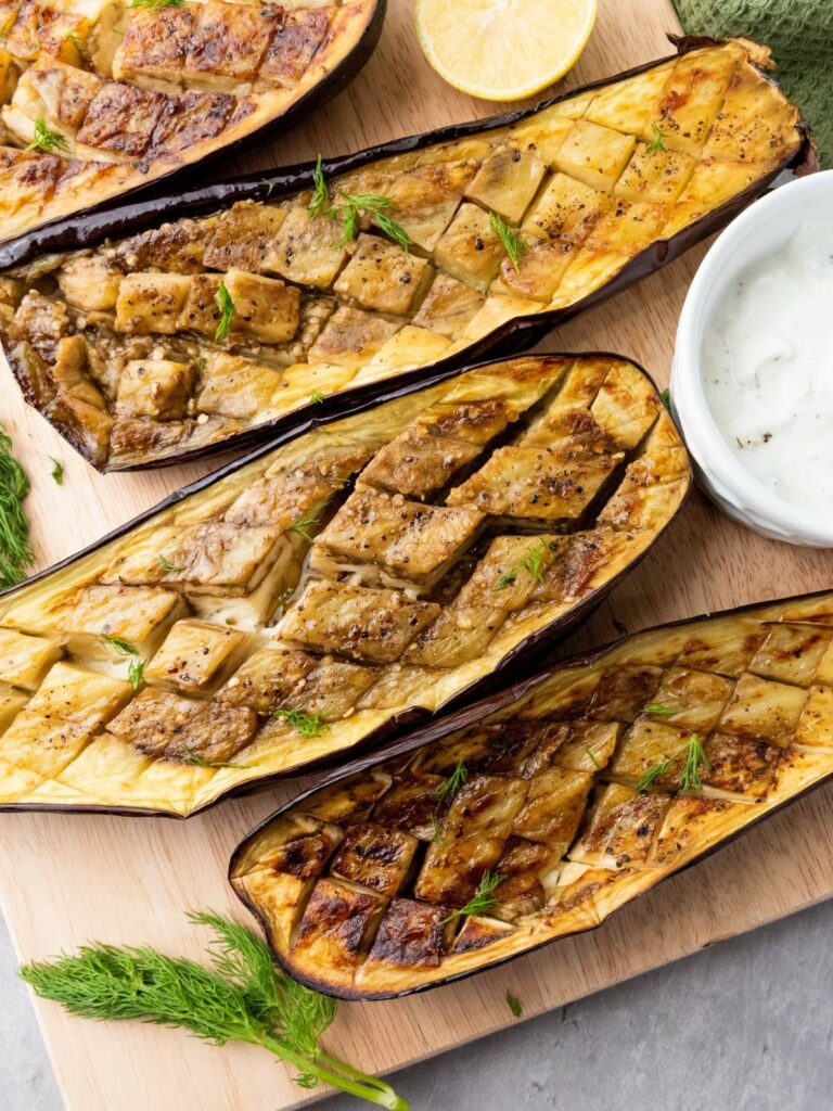 Four roasted eggplant halves with a scored pattern are arranged on a wooden board.