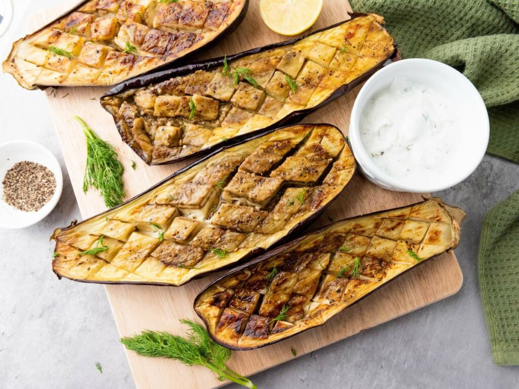 Four roasted eggplant halves with a crisscross pattern served on a wooden board.