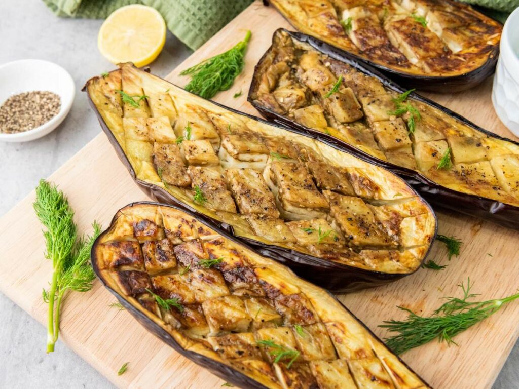 Roasted eggplant halves with scored flesh are arranged on a wooden board, garnished with fresh dill.