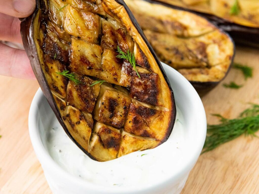 A roasted, scored eggplant half is being dipped into a bowl of creamy white sauce.
