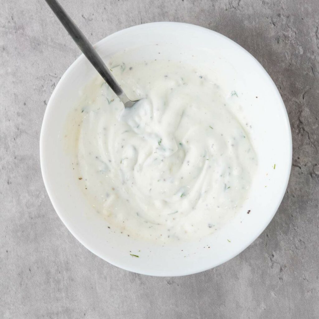 A white bowl filled with creamy white sauce, speckled with herbs and pepper, with a spoon resting inside.