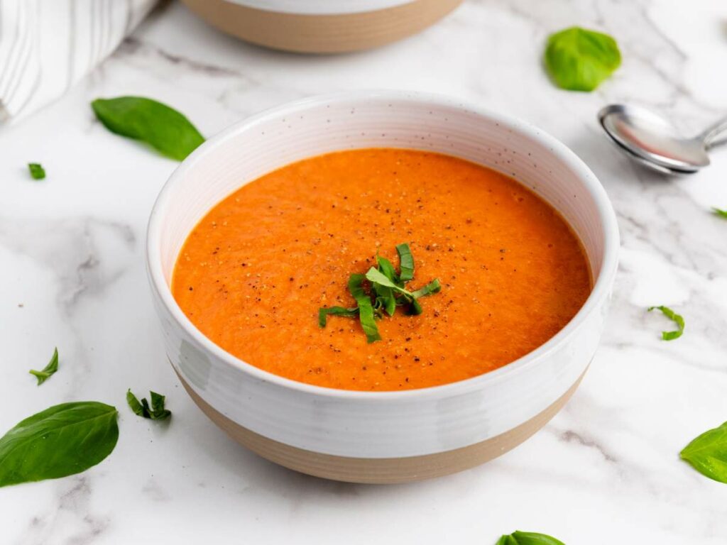 A bowl of tomato soup garnished with chopped basil sits on a marble surface, with a spoon and basil leaves nearby.