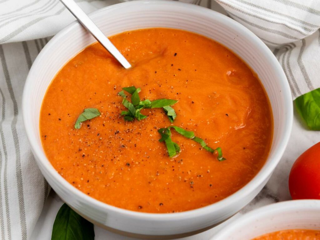 A bowl of tomato soup garnished with chopped fresh basil and black pepper, with a spoon resting in the bowl.