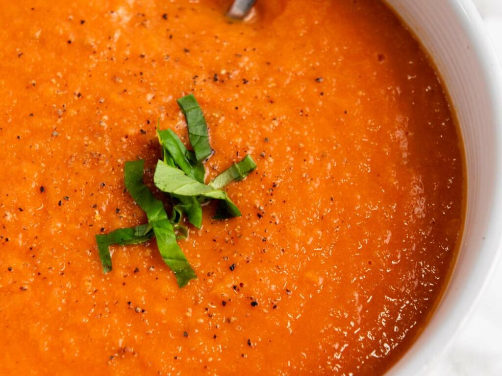 A close-up of a bowl of tomato soup, topped with ground black pepper and chopped basil.