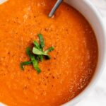 A bowl of tomato soup garnished with chopped basil and black pepper, with a spoon in the bowl.