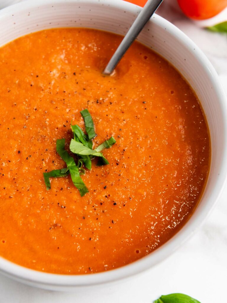 A bowl of tomato soup garnished with chopped basil and black pepper, with a spoon in the bowl.