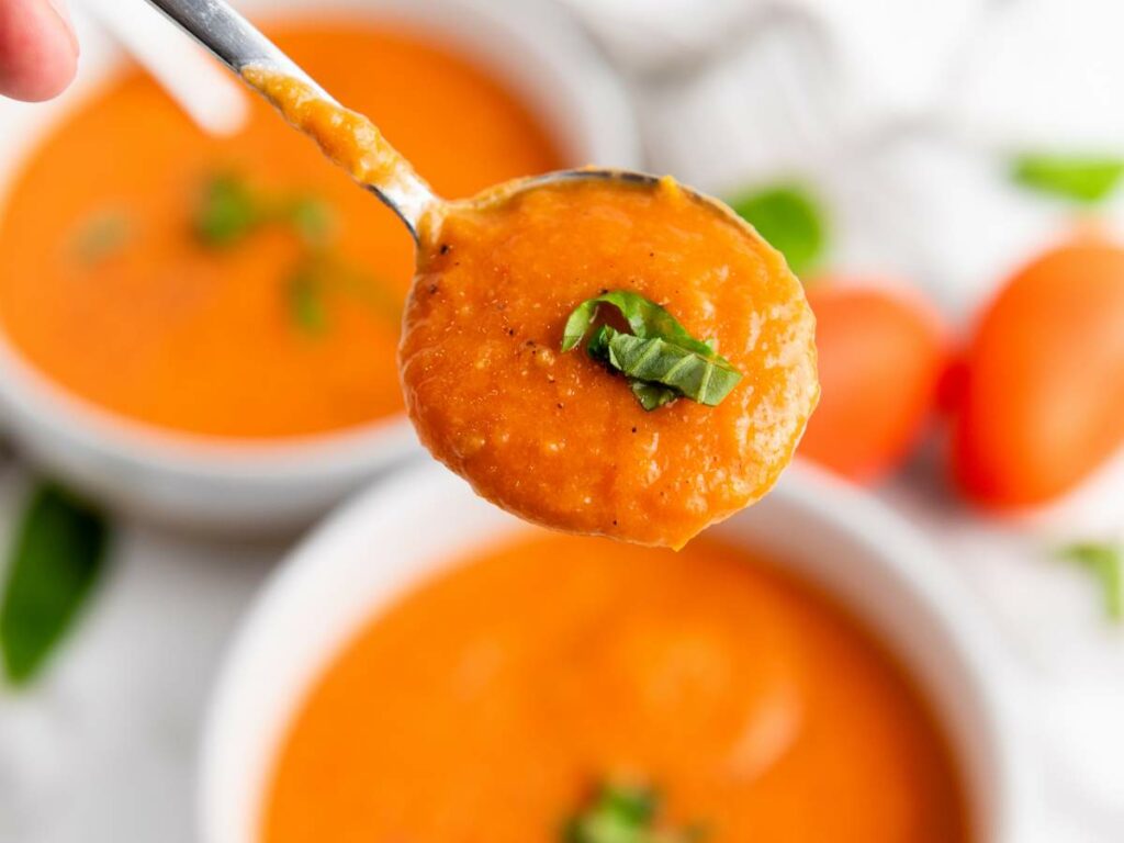 A spoonful of creamy tomato soup garnished with fresh basil, held above a bowl of similar soup.