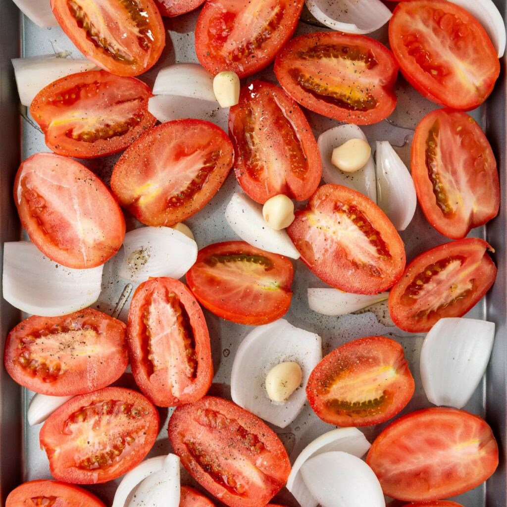 Halved tomatoes, quartered onions, and whole garlic cloves arranged on a baking tray, seasoned with olive oil, salt and pepper.
