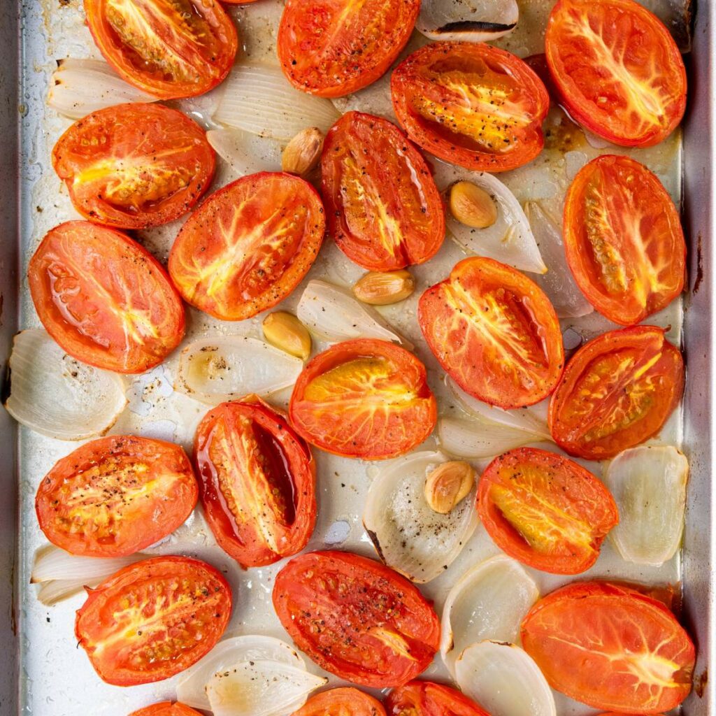 Halved roasted tomatoes, onion wedges, and a few whole garlic cloves on a baking sheet.