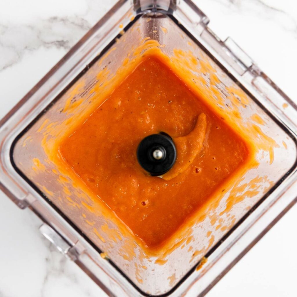 A blender viewed from above contains a smooth tomato soup on a white marble surface.