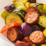 Sausage, zucchini, and peppers are beautifully baked with a sprinkle of herbs in a white bowl.