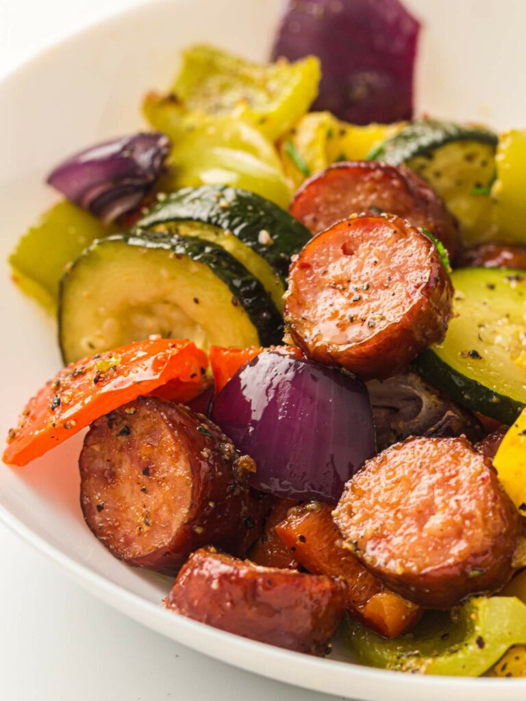 Sausage, zucchini, and peppers are beautifully baked with a sprinkle of herbs in a white bowl.