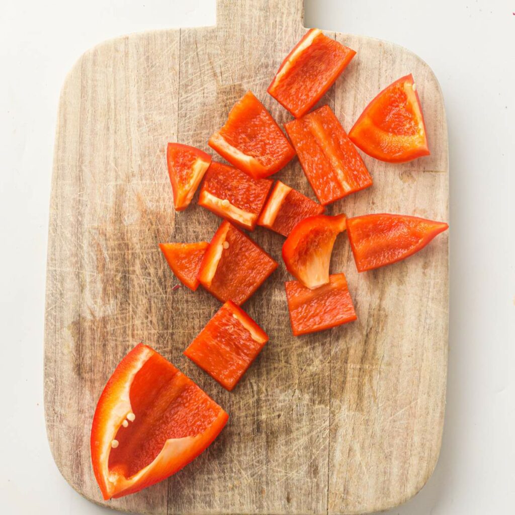 Chopped red bell pepper pieces are spread out on a wooden cutting board.