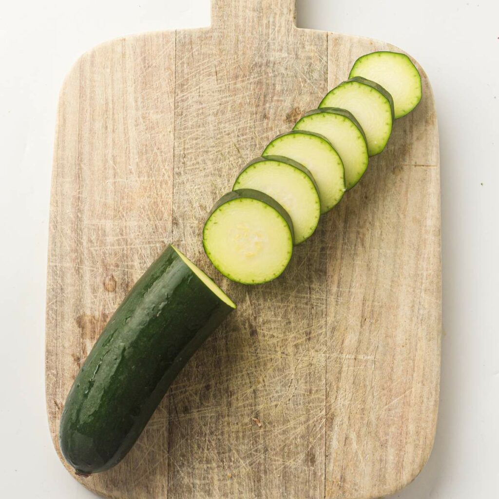 A zucchini is partially sliced into even rounds on a wooden cutting board.