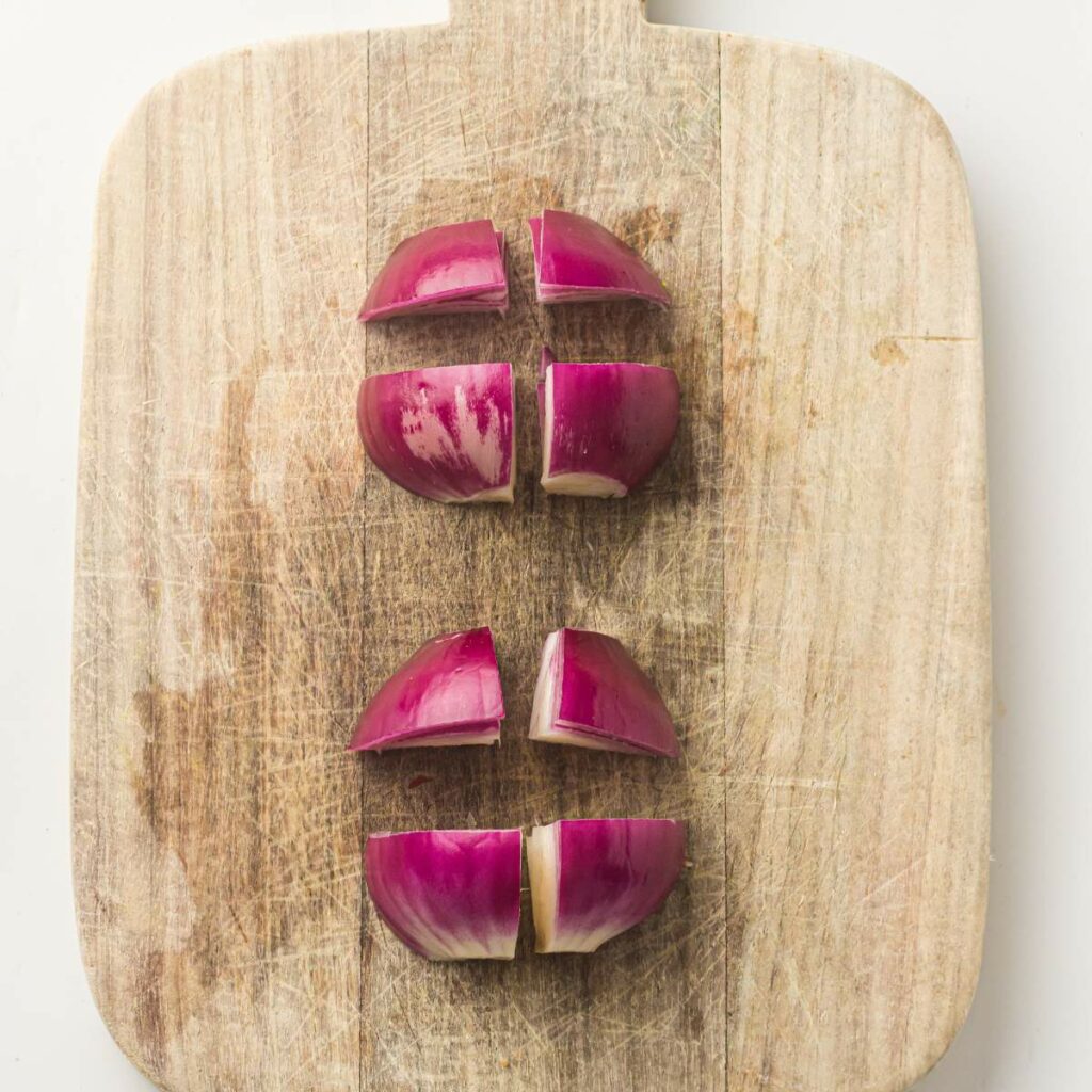 Red onion is cut into wedges and placed on a wooden cutting board. A small glass bowl of olive oil is held above a tray of fresh vegetables.