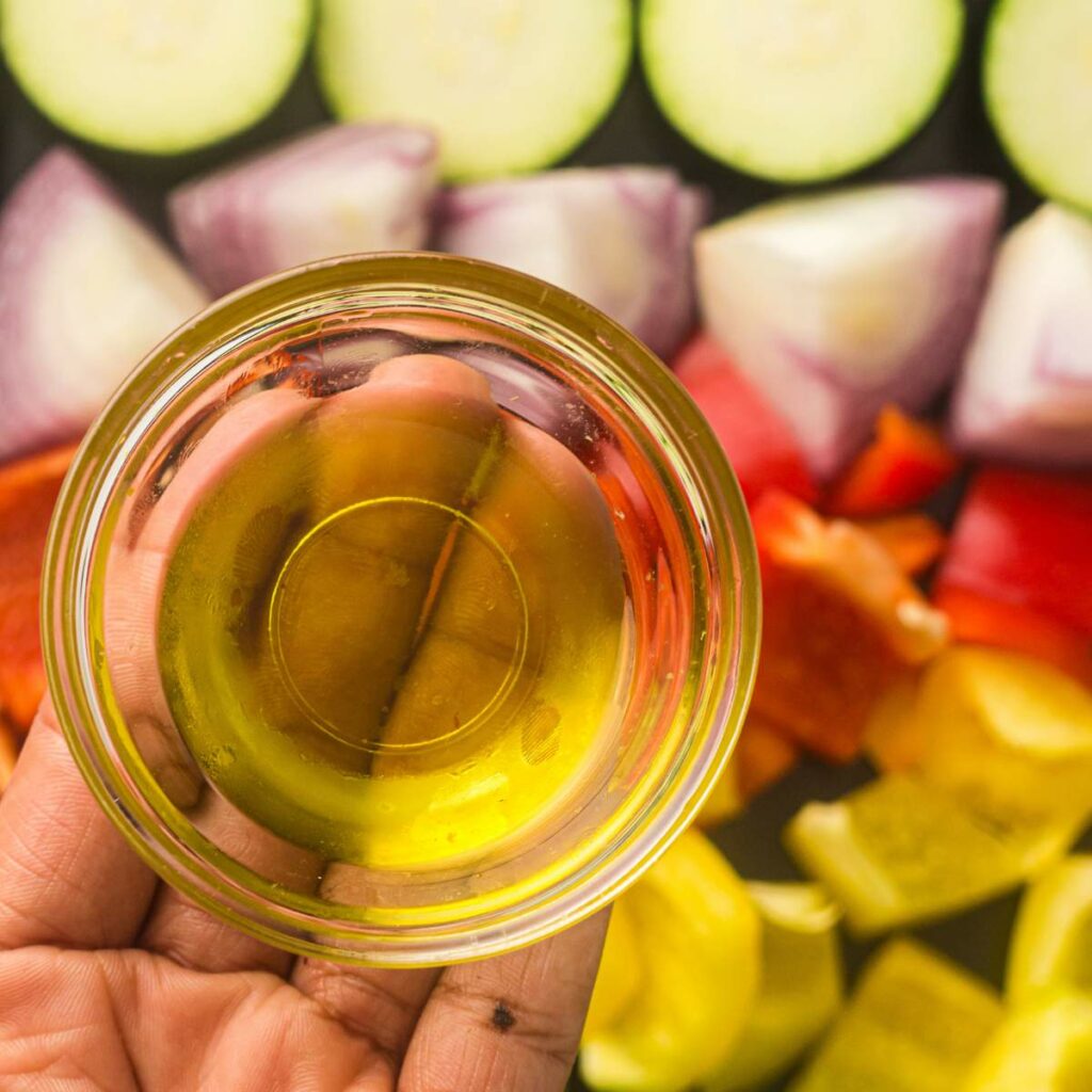 A small glass bowl of olive oil is held above a tray of fresh vegetables.