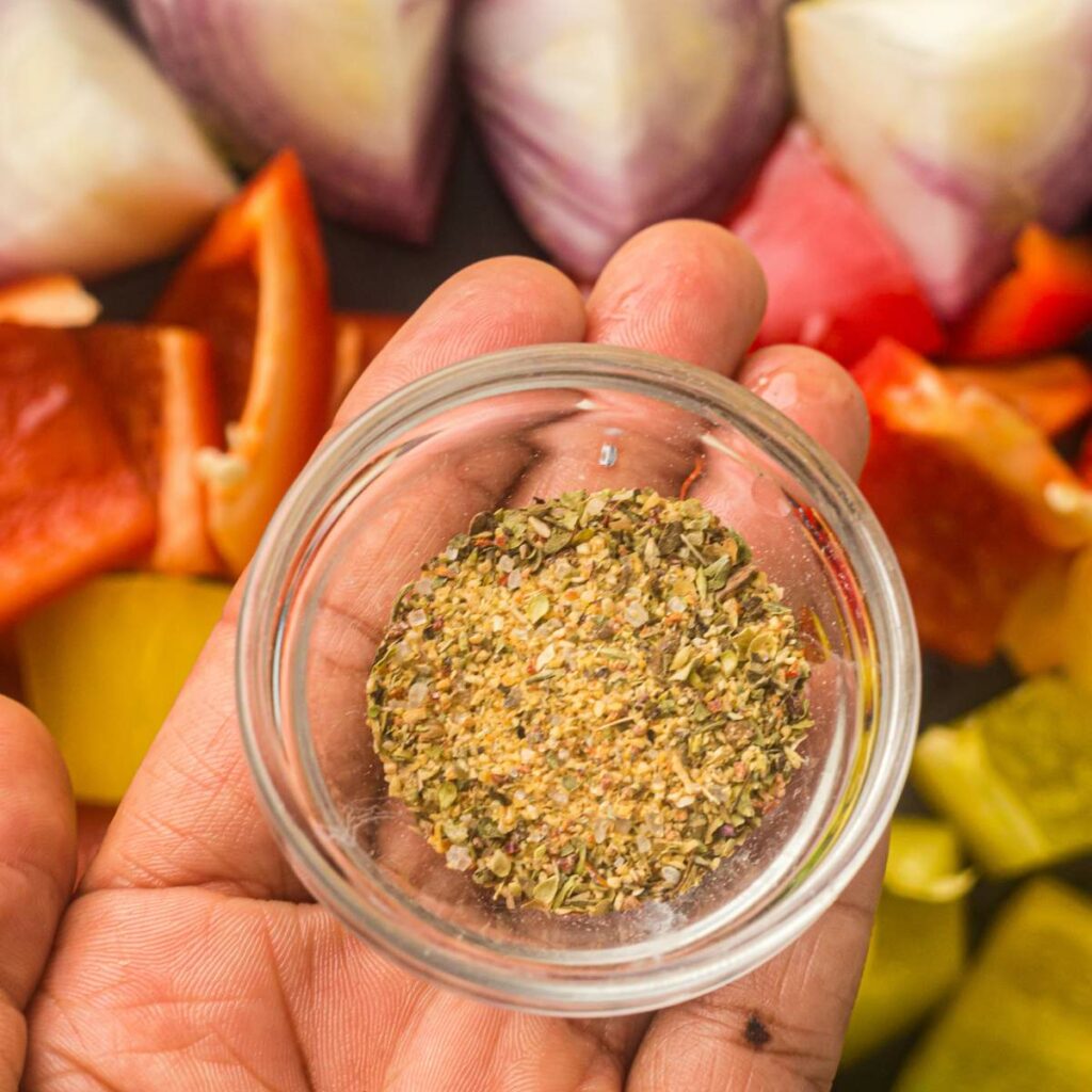 A small glass bowl of Italian seasoning is held above a tray of vegetables.