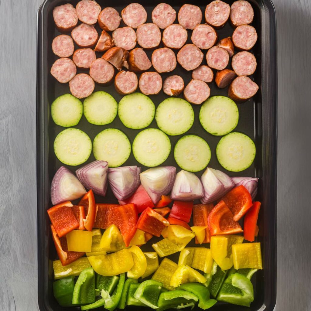 A baking sheet is neatly lined with rows of sausage, zucchini, onion, and bell peppers.