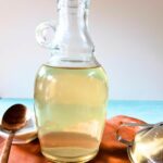 Clear glass bottle filled with a transparent liquid, place on a wooden tray next to a metal spoon in the background.