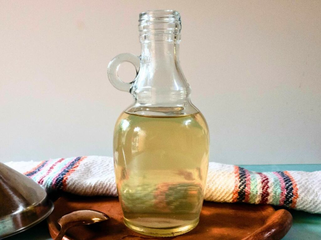 A clear glass bottle filled with a light golden liquid is placed on a wooden tray with a striped white towel in the background.