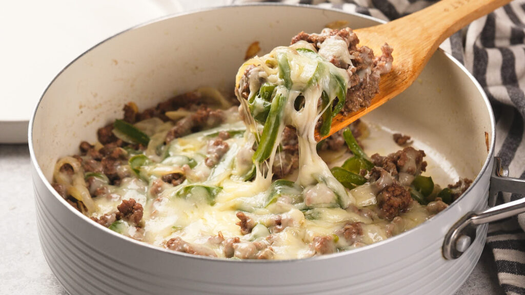 Ground beef Philly cheesesteak Skillet with a wooden spoon lifting a portion of the cheesy mixture.