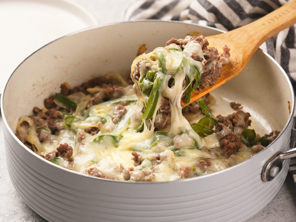 A skillet filled with cooked Philly cheesesteak being scooped with a wooden spoon.