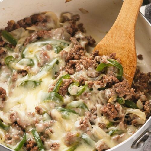 Ground beef, melted cheese, sliced green bell peppers, and onions are mixed together in a white pan with a wooden spoon.