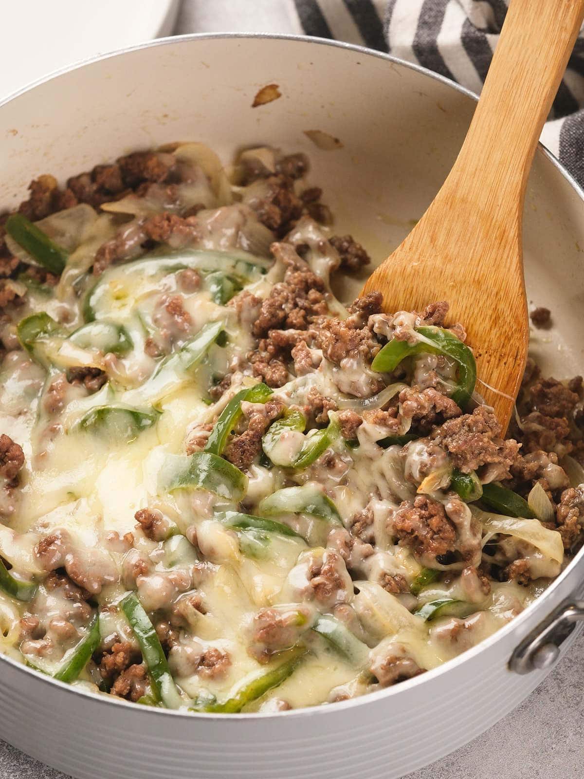 Ground beef, melted cheese, sliced green bell peppers, and onions are mixed together in a white pan with a wooden spoon.