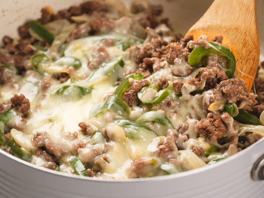 Ground beef philly cheesesteak in a white skillet with a wooden spatula.