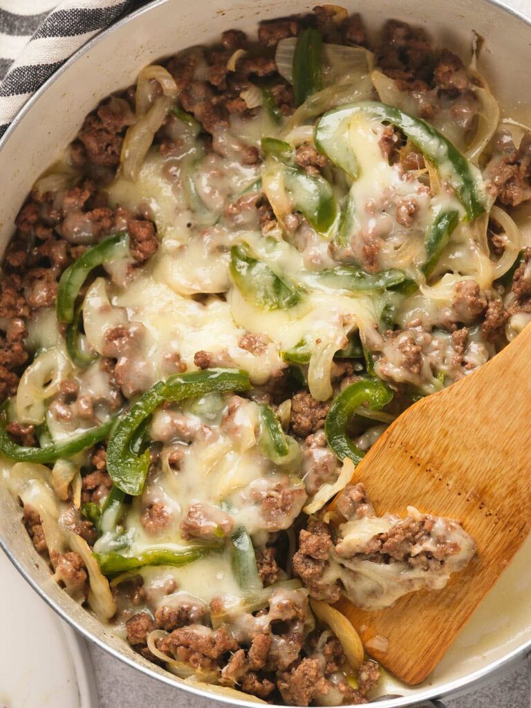 A skillet filled with cooked ground beef, sliced green bell peppers, onions, and melted cheese, being stirred with a wooden spatula.