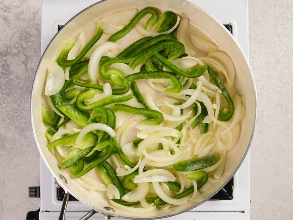 Sliced green bell peppers and onions cooking in a pan on a stovetop.