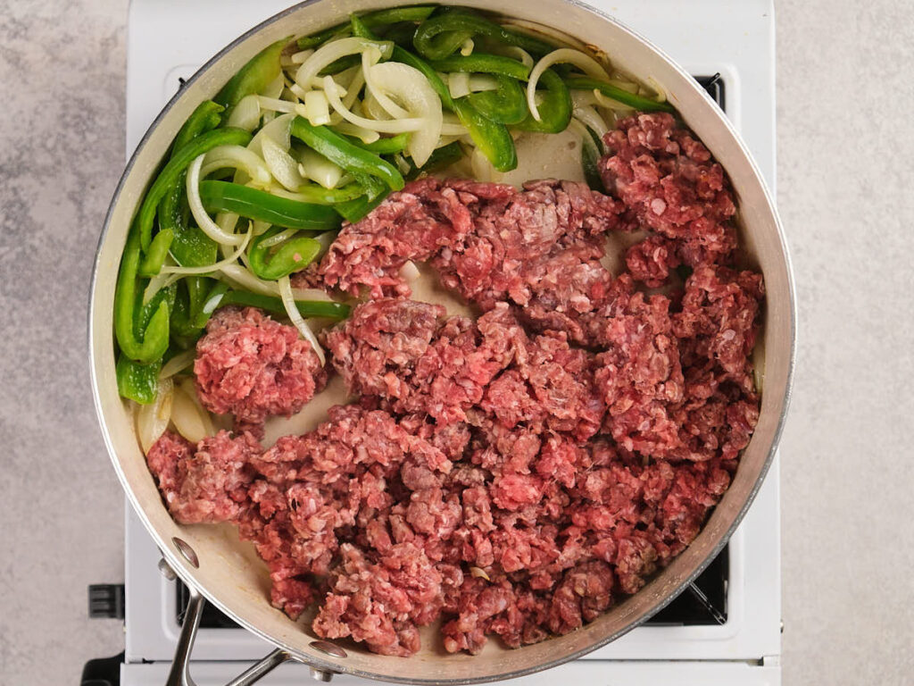 Ground beef, sliced green bell peppers, and onions cooking in a skillet on a stovetop.