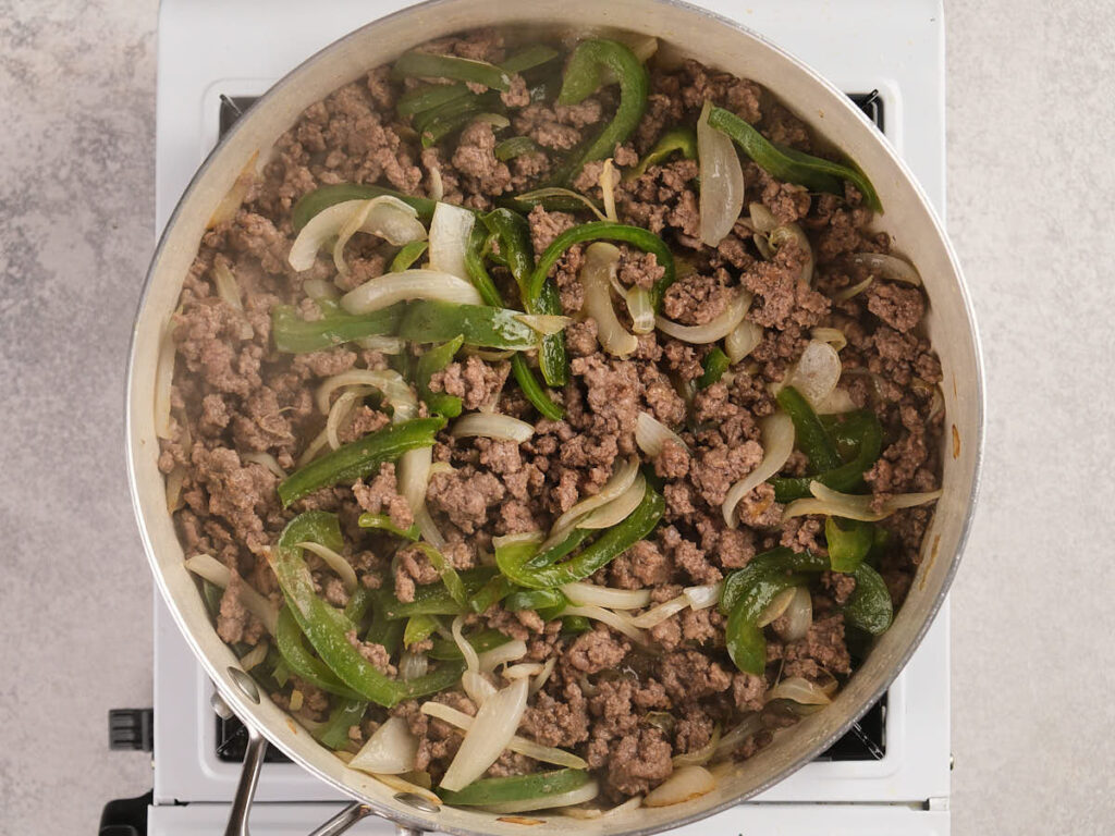 Ground beef, sliced green bell peppers, and onions are cooking in a large skillet on a stovetop.