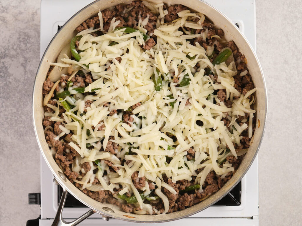 A skillet filled with cooked ground beef, green bell peppers, and onions, topped with a layer of shredded cheese.