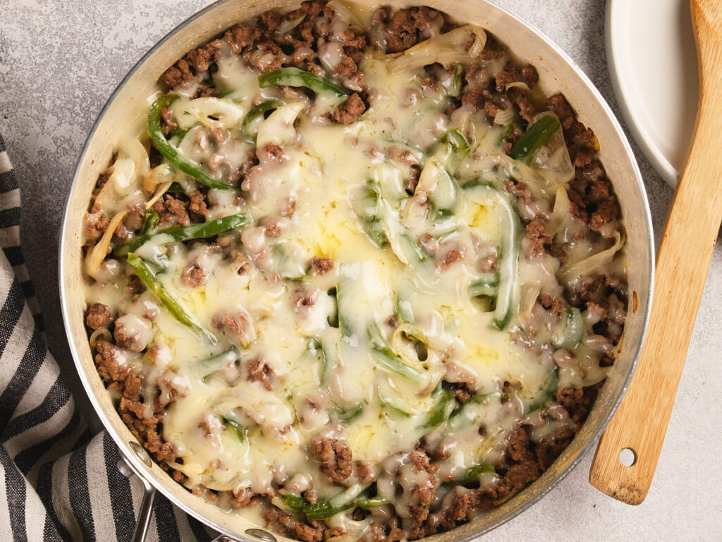 A skillet filled with cooked ground beef, sliced green bell peppers, onions, and melted cheese on top, with a wooden spoon beside it.