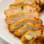 Sliced cooked chicken breast arranged on a white plate, garnished with chopped herbs.