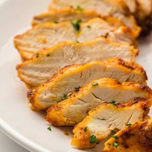 Sliced cooked chicken breast arranged on a white plate, garnished with chopped herbs.