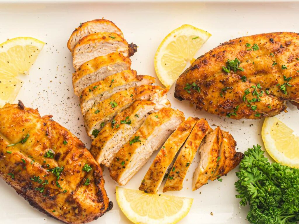 Cooked chicken breasts, some sliced, garnished with chopped herbs and served with lemon wedges and parsley on a white plate.