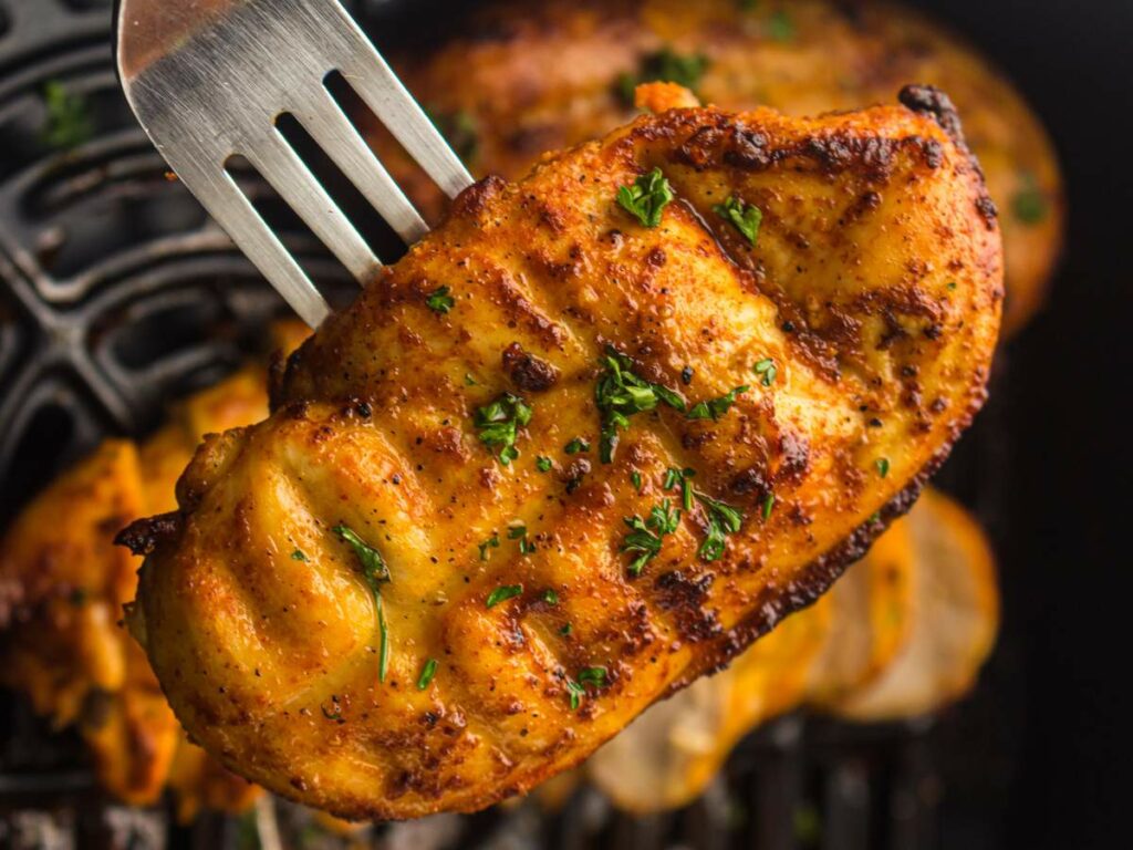 A cooked, seasoned chicken breast garnished with chopped parsley is held up by a metal fork over an air fryer basket.