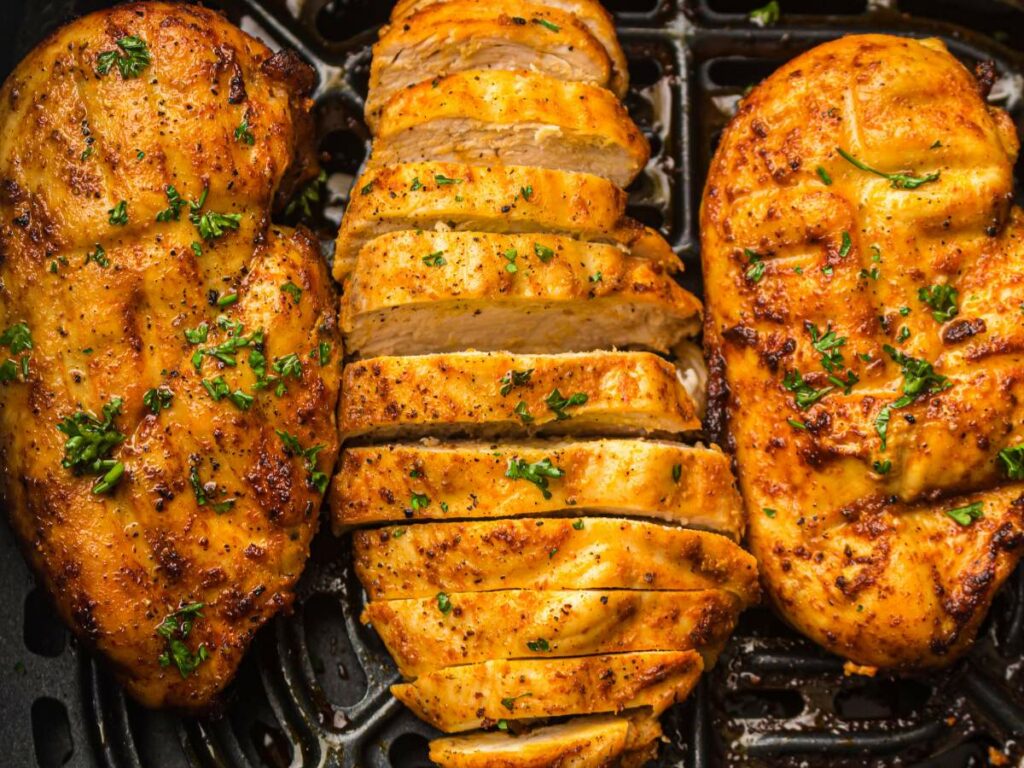 Three seasoned chicken breasts, two whole and one sliced, cooked in an air fryer basket and garnished with chopped herbs.