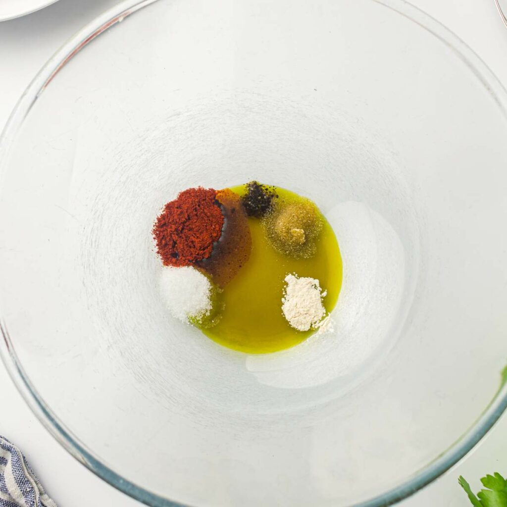 A glass bowl containing olive oil, paprika, salt, pepper, garlic powder, and onion powder, viewed from above.