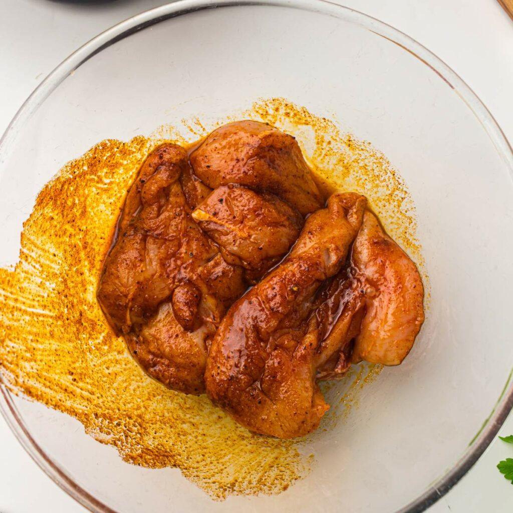 Raw chicken breasts coated in a reddish-brown marinade sit in a clear glass bowl on a white surface.